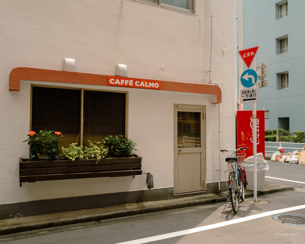 A small café called Caffè Calmo on a quiet street corner in Tokyo, with a bicycle parked outside and a Coca-Cola vending machine nearby.