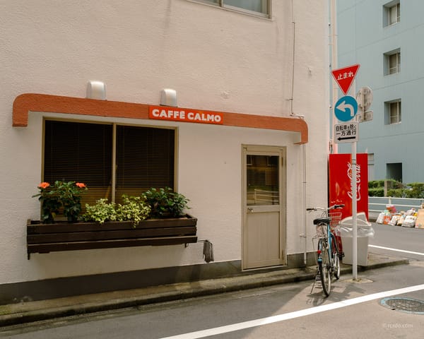 A small café called Caffè Calmo on a quiet street corner in Tokyo, with a bicycle parked outside and a Coca-Cola vending machine nearby.